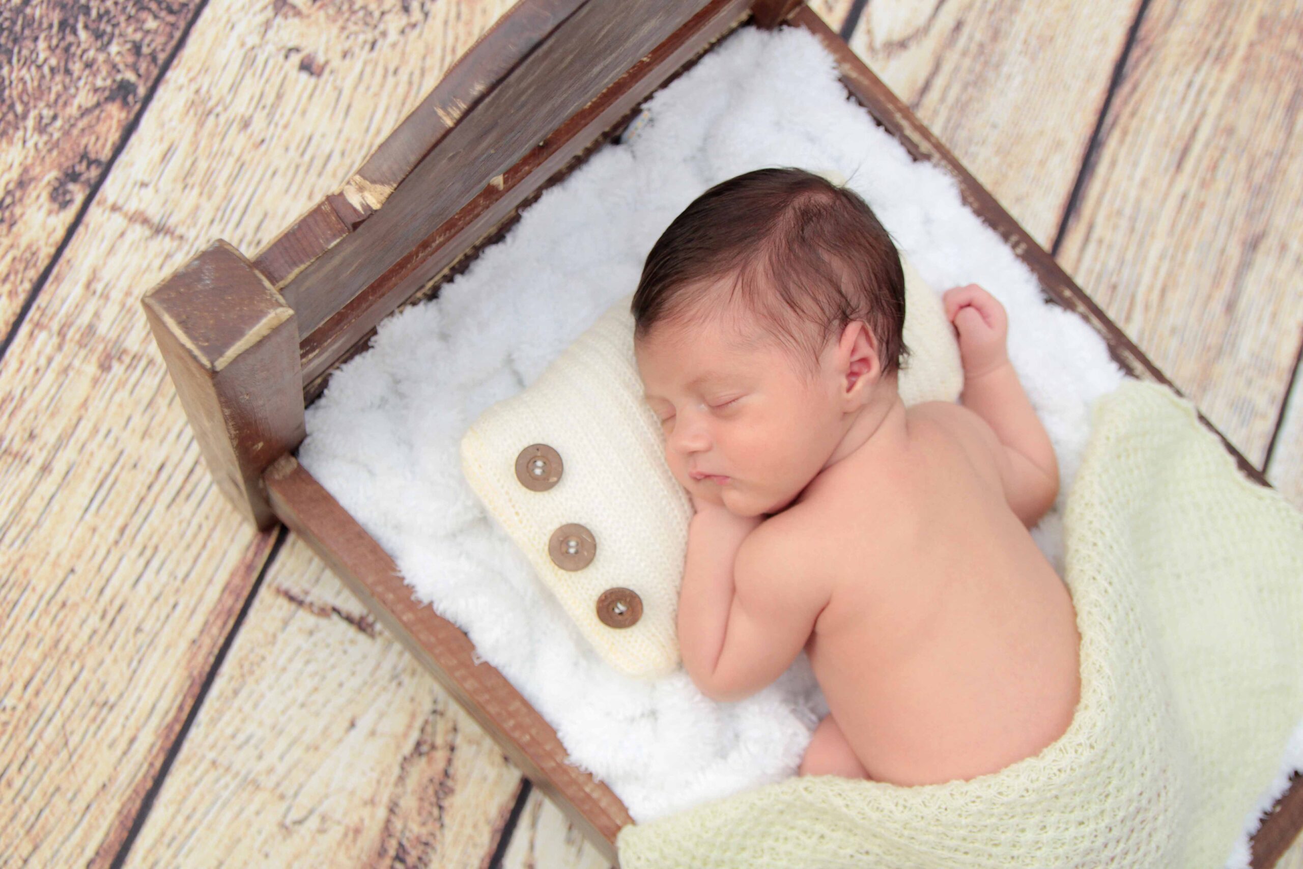 Bebê recém-nascido dormindo de lado em mini cama rústica com cobertor claro, durante ensaio newborn feito por Estefani Fontes.